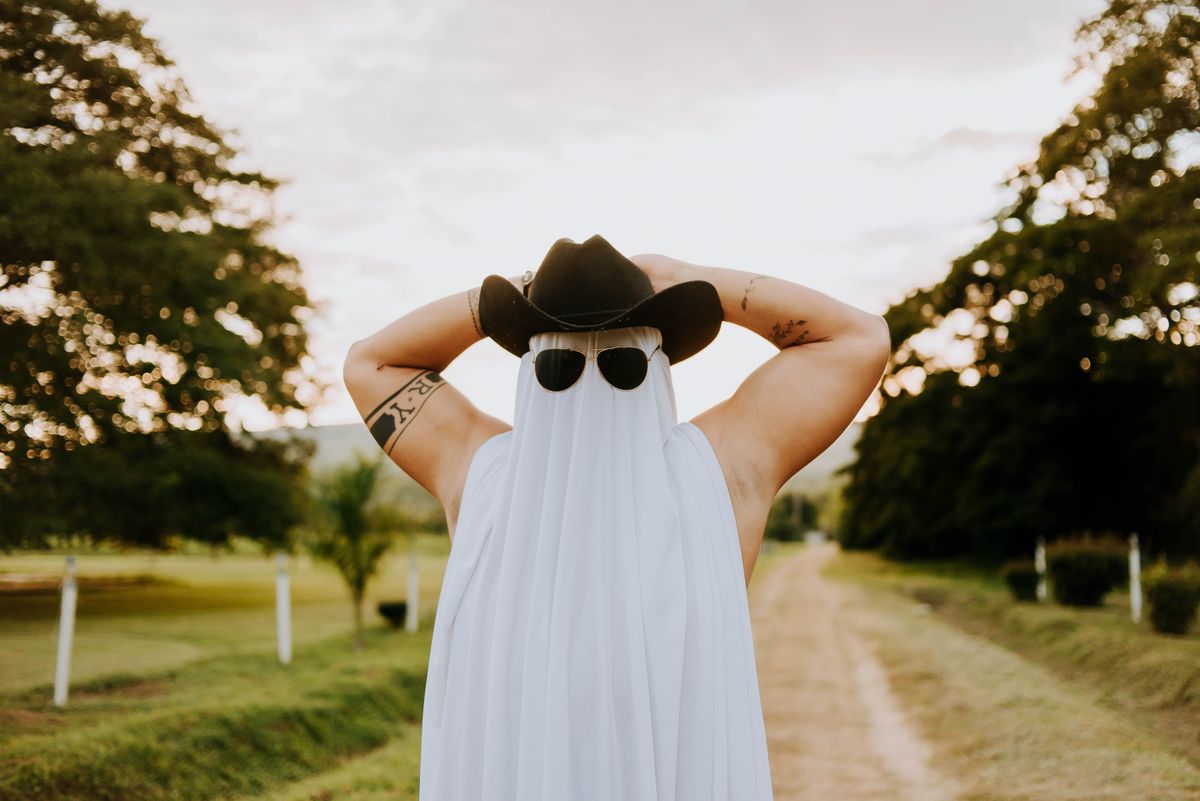 Muscled man posing as a ghost in cowboy hat and sunglasses on a rural road.