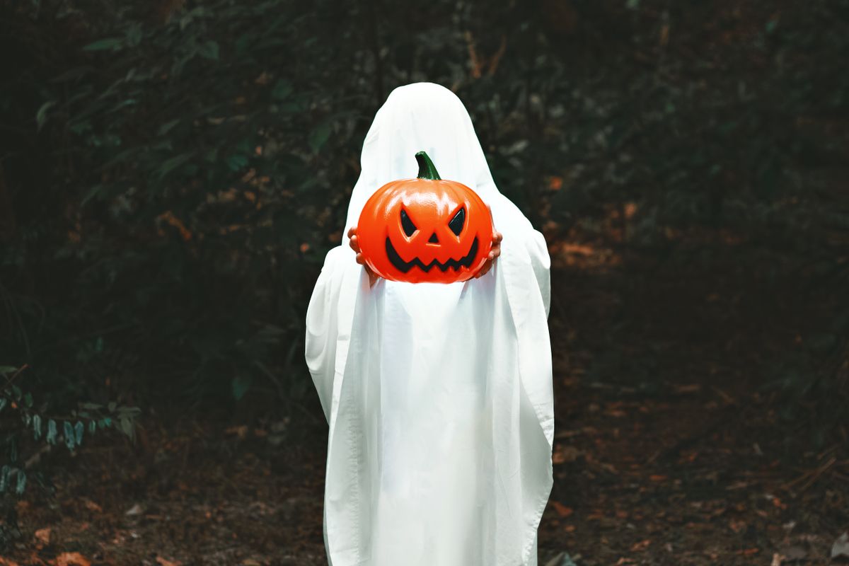 A person in a ghost costume holds a Jack-o'-lantern in a dark, eerie forest setting.