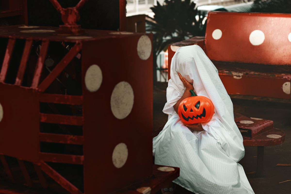 A person dressed as a ghost holding a jack-o'-lantern, sitting on a red and white bench.