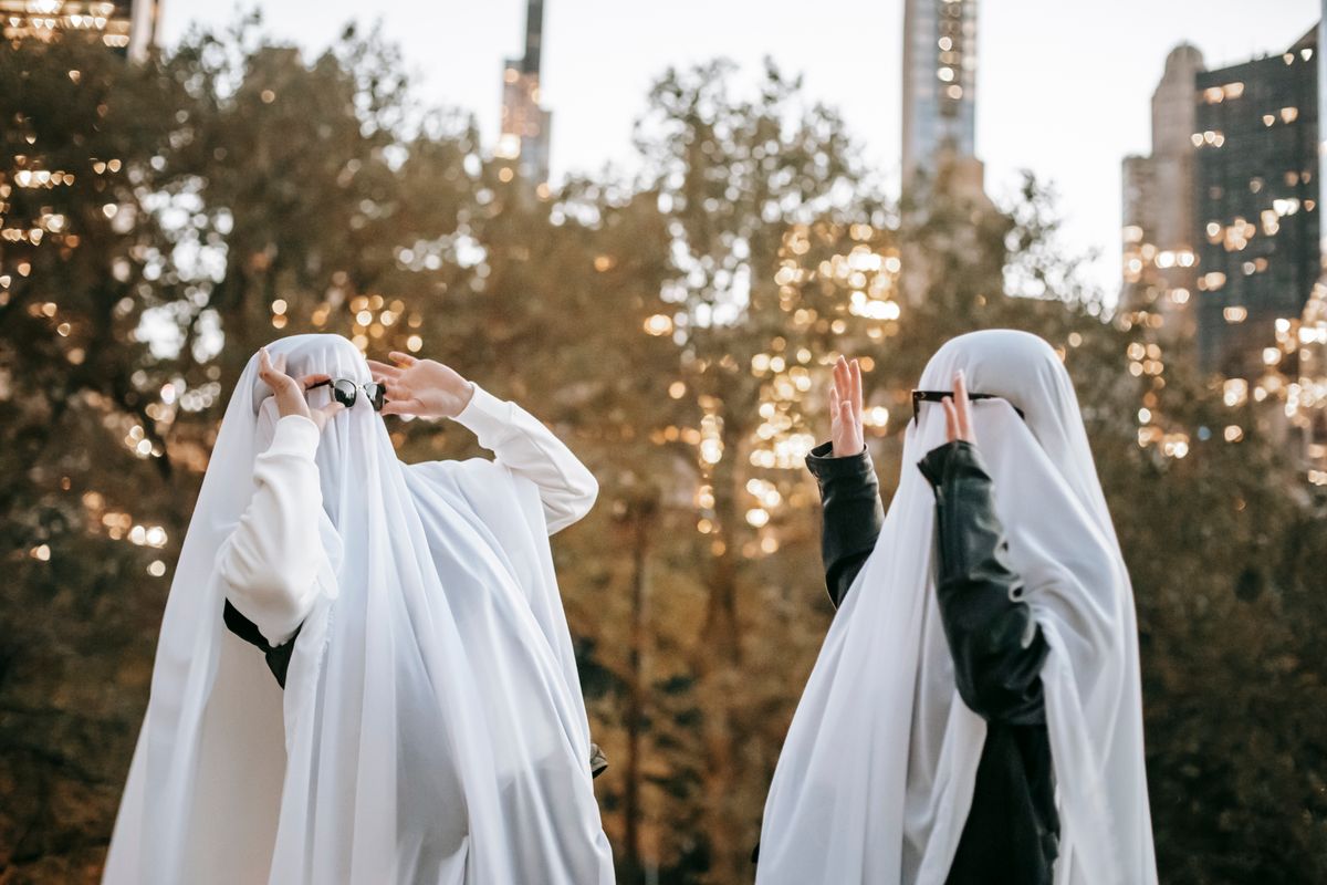 Side view of spooky unrecognizable couple with raised arms wearing ghost costumes and sunglasses looking at each other while standing on street with trees on blurred background during Halloween