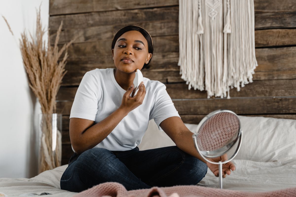 Woman practicing skincare routine at home using a facial massager while sitting on bed.