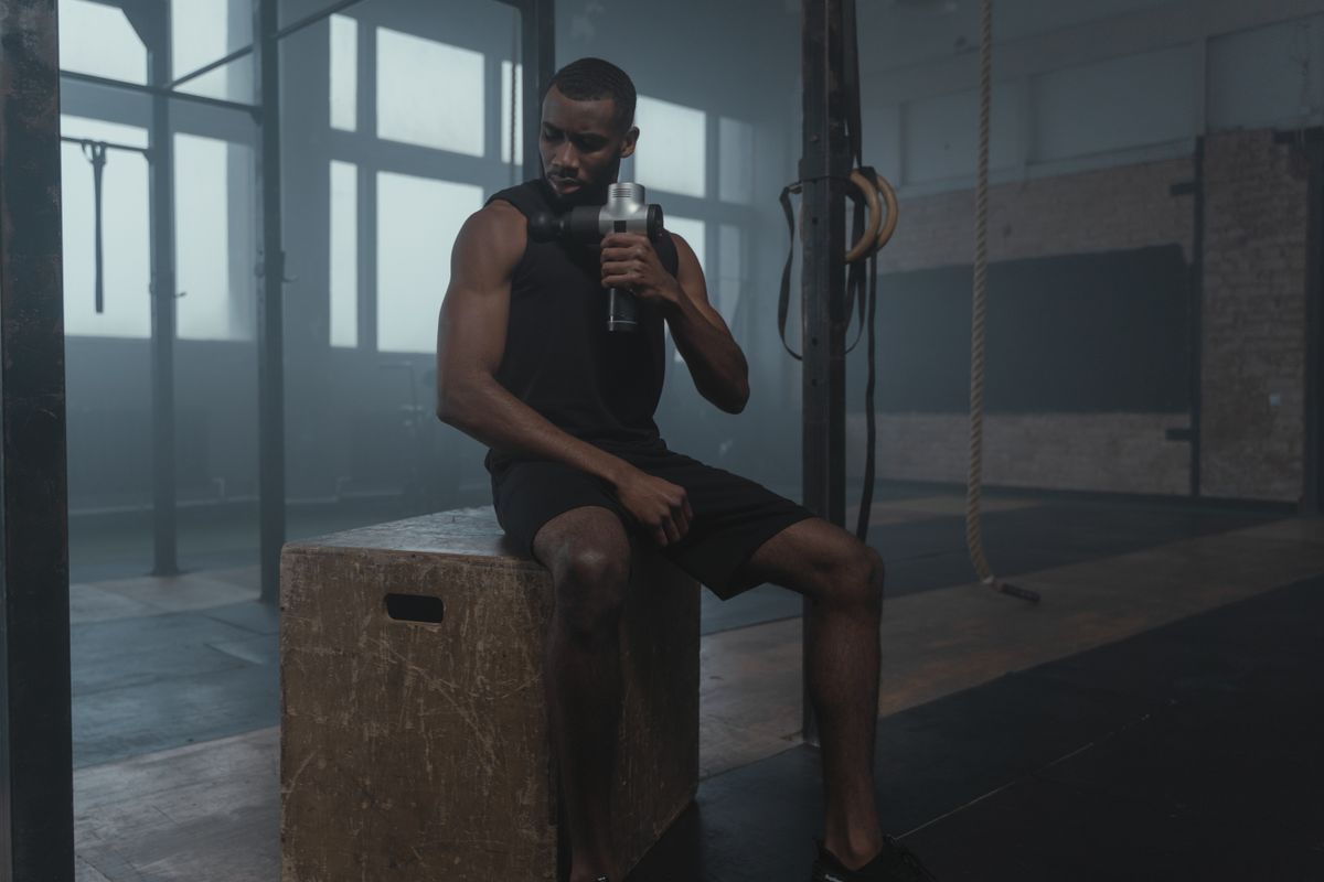 Fit man using a massage gun in a gym, wearing a tank top while seated on a wooden box.