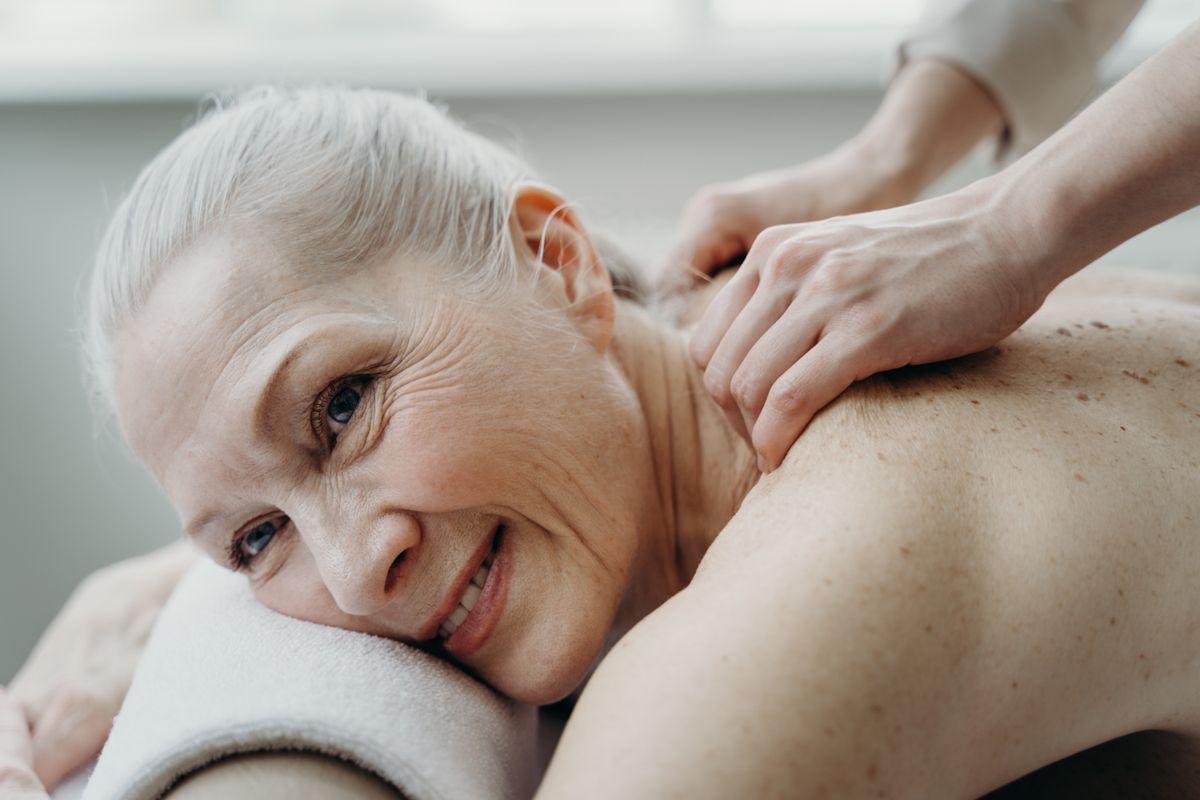 A senior woman with white hair smiles while receiving a therapeutic back massage indoors.