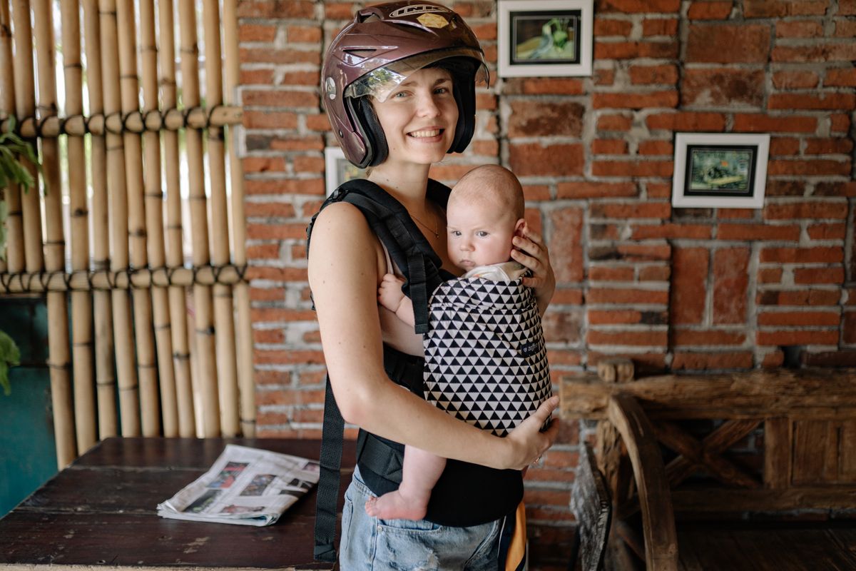 A smiling mother holding her baby in a carrier, both indoors with a helmet on.