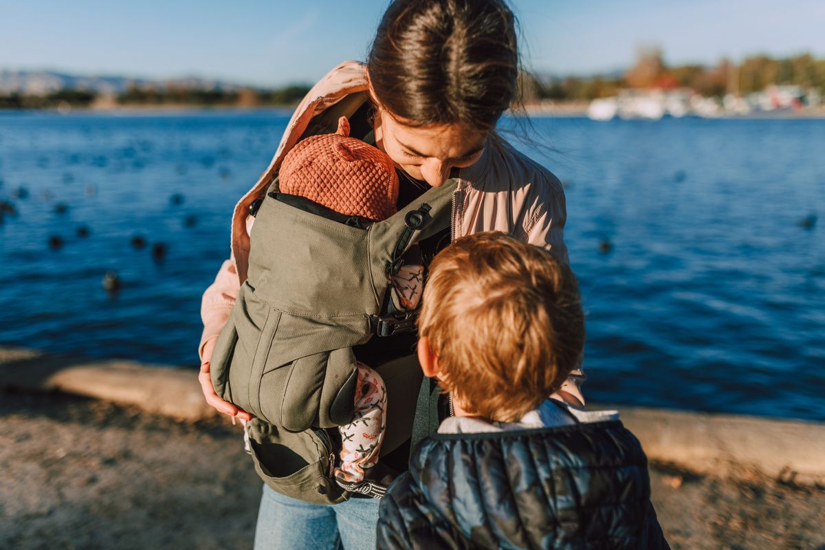 A mother holding her baby in a carrier while her young son stands nearby by a scenic waterfront.
