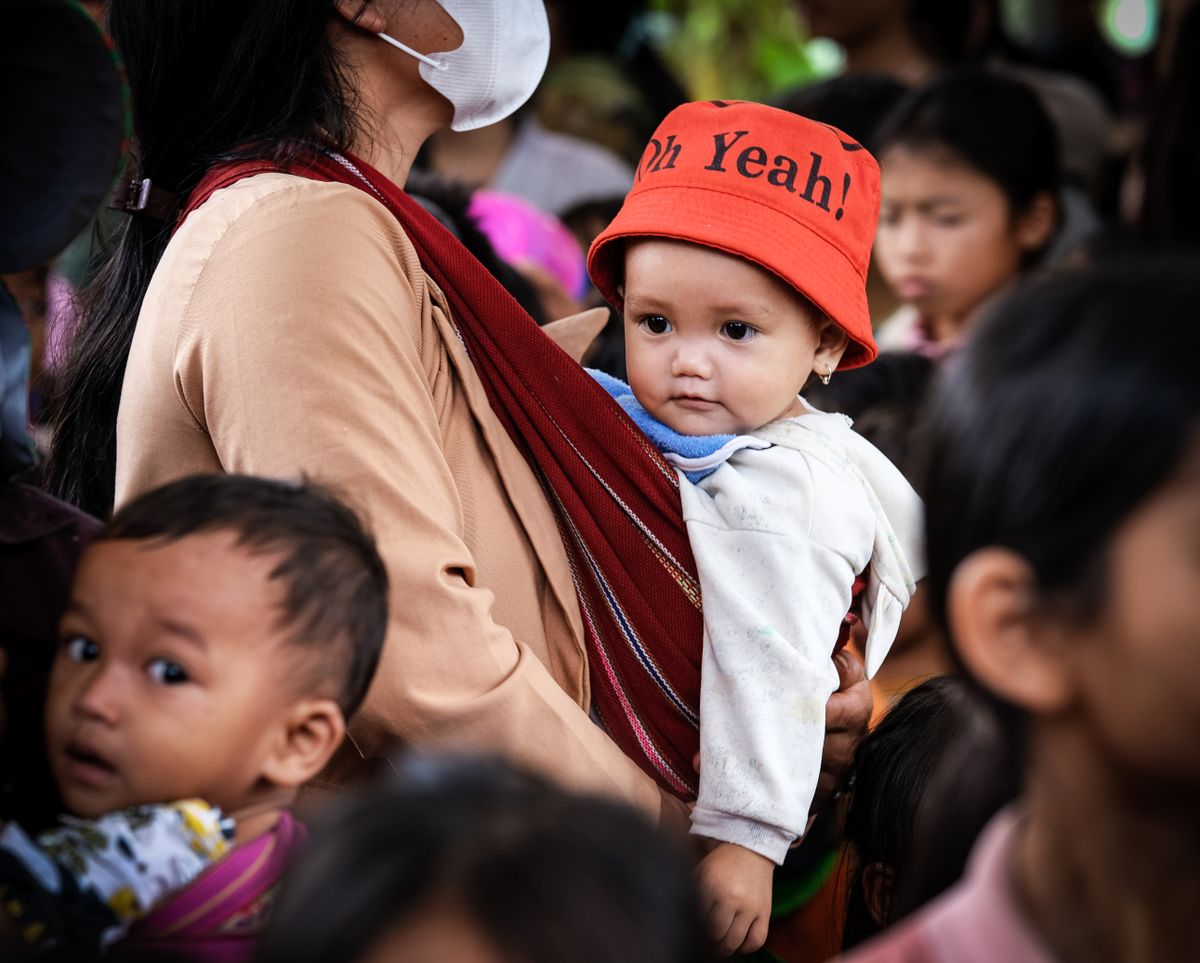 A mother carrying her baby with a bold hat amidst a lively crowd. Captured outdoors.