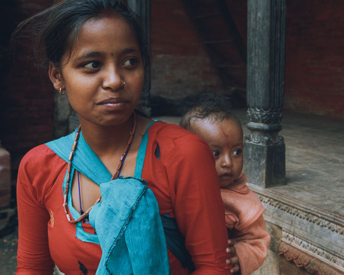 A young mother carries her baby, wrapped in traditional attire, in a rustic setting.