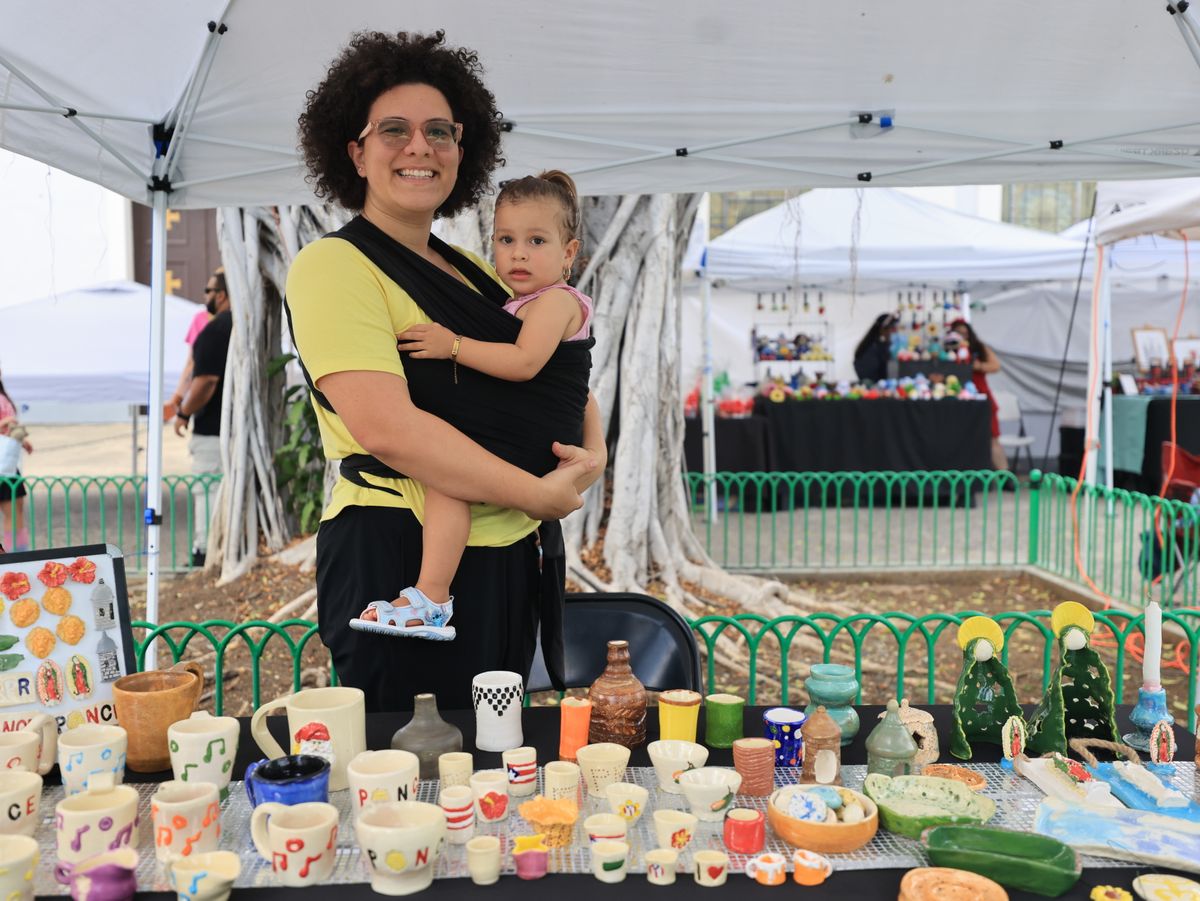 A mother holding her child at an outdoor pottery market stall showcasing colorful handmade ceramics.