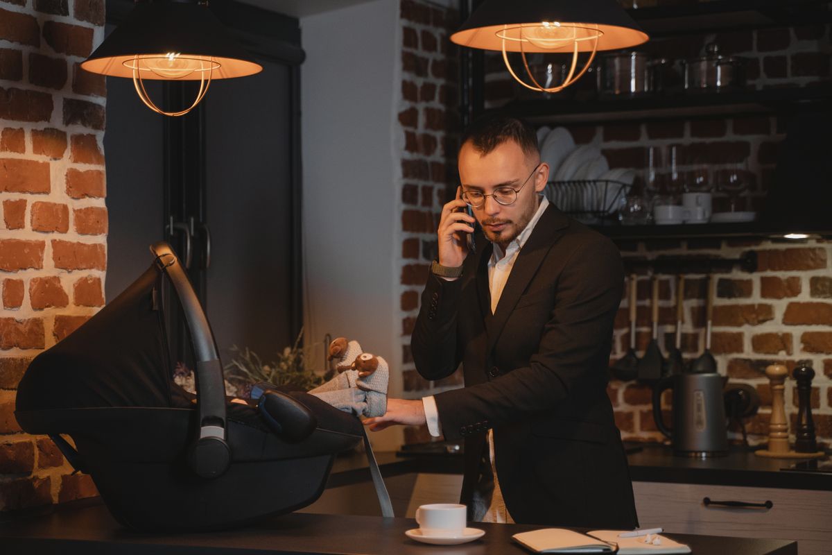 Man in suit multitasks with baby in carrier while on a phone call in stylish kitchen.