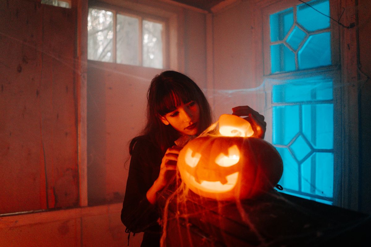 A woman carves a glowing jack-o'-lantern indoors amidst cobwebs and eerie lighting, celebrating Halloween.