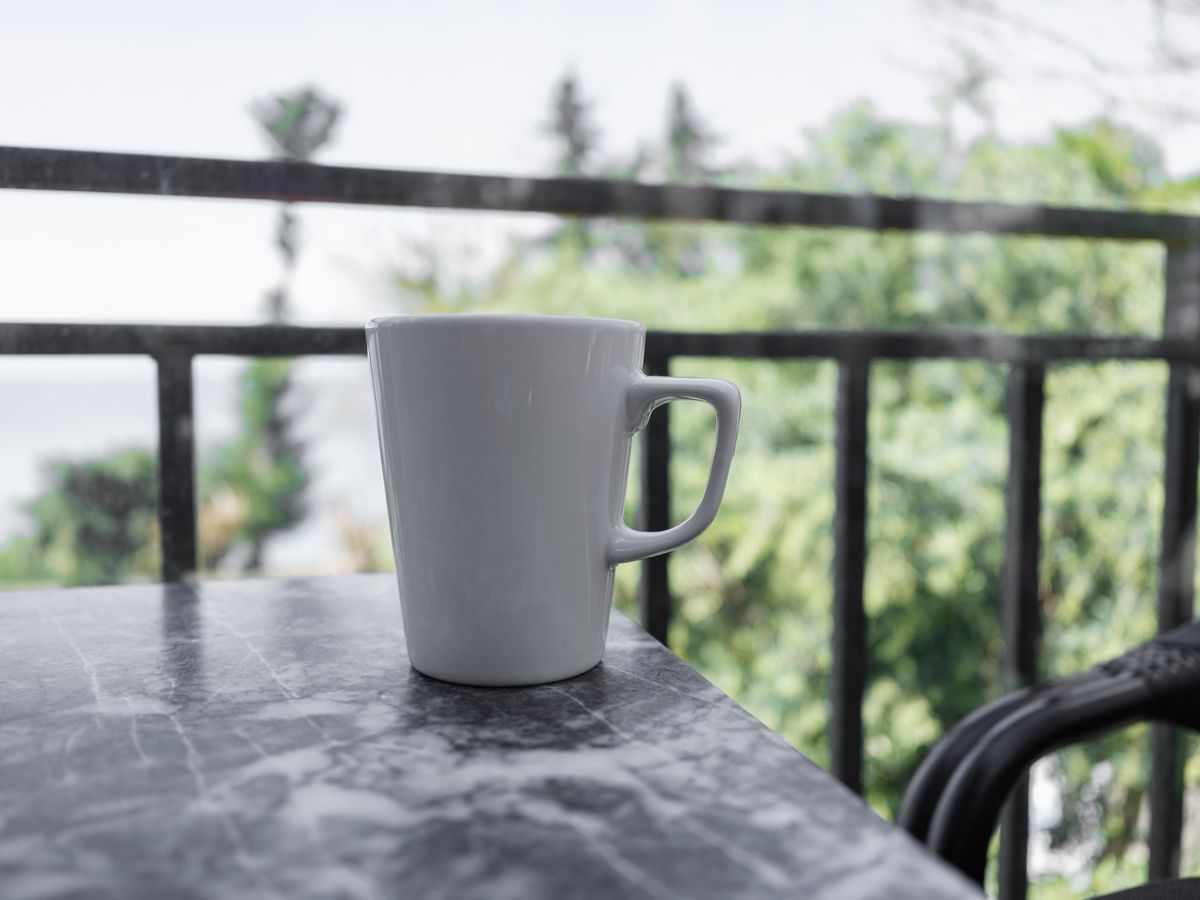 A white mug of coffee sits on a marble table with a scenic outdoor view of greenery.