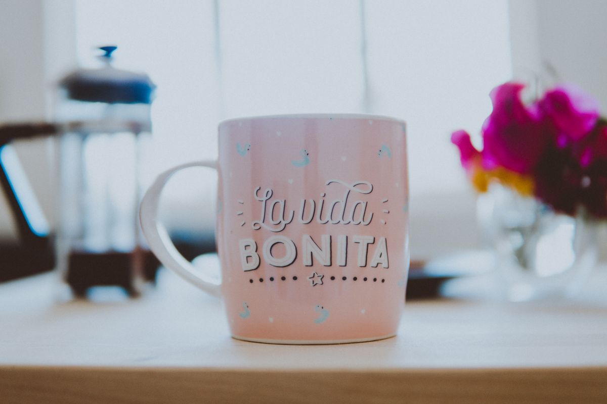 A pink mug with 'La vida BONITA' text, on a table with flowers and a French press.