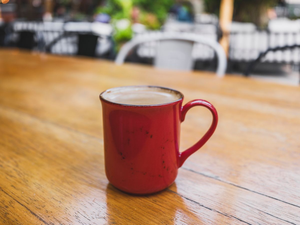 A steaming hot coffee served in a vibrant red ceramic mug placed on a rustic wooden table outdoors.