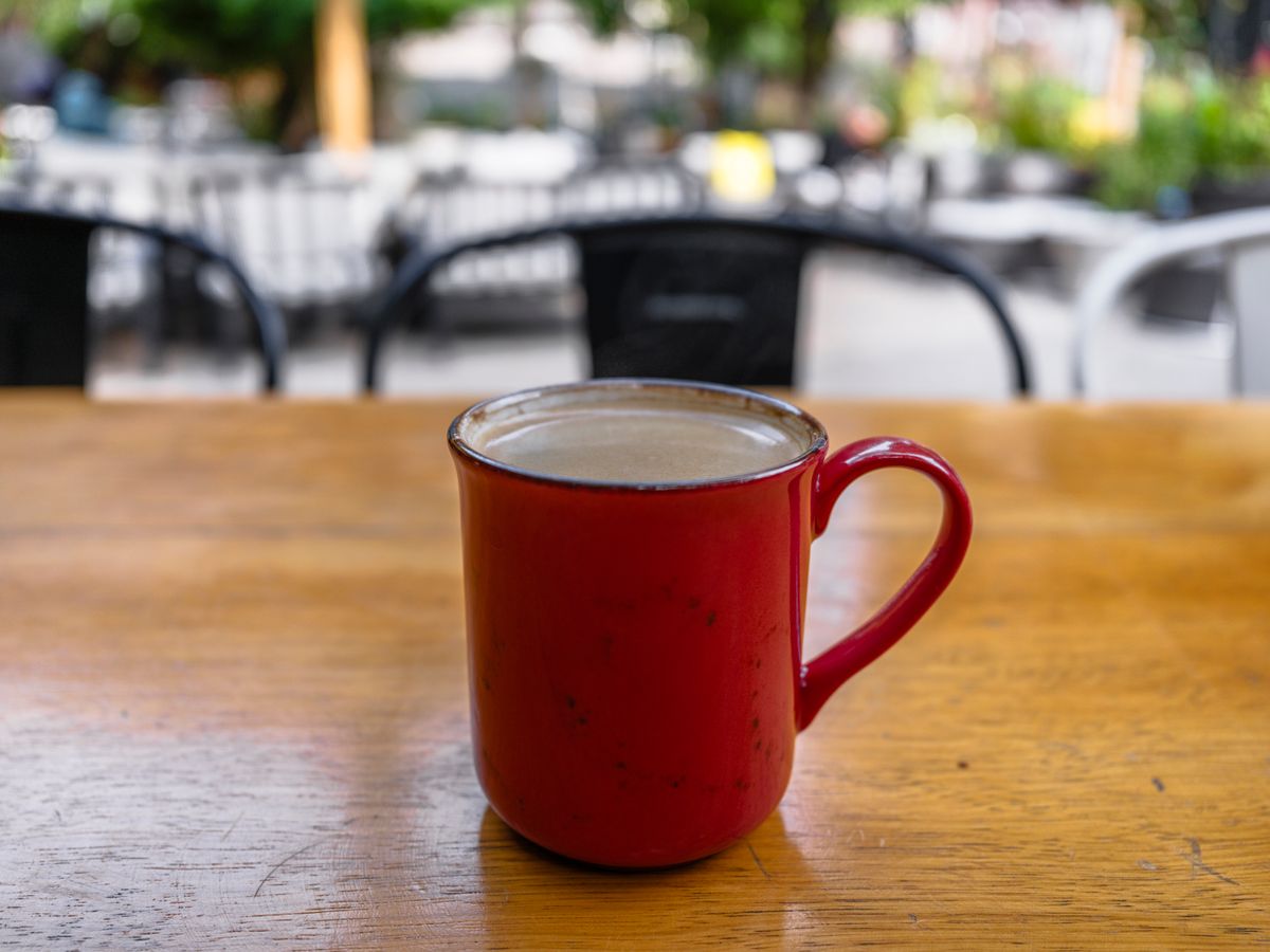 A rustic red ceramic mug filled with hot coffee on a wooden table in an outdoor café setting.