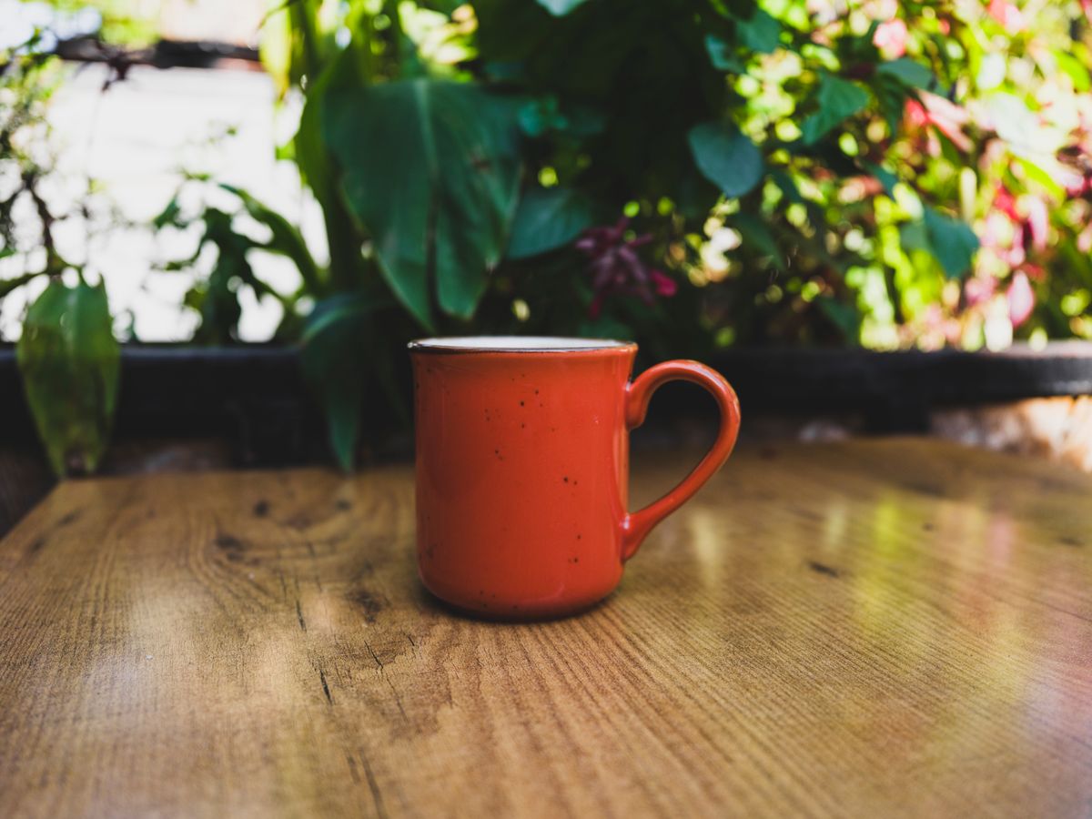 Red mug on a wooden table outdoors with vibrant greenery in the background, suggesting a cozy setting.