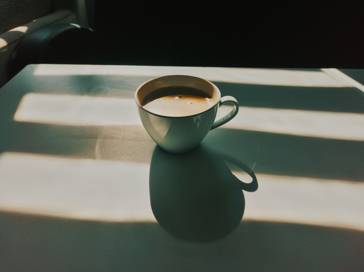 A white coffee cup sits on a table with striking sunlight and shadows creating a warm ambiance.