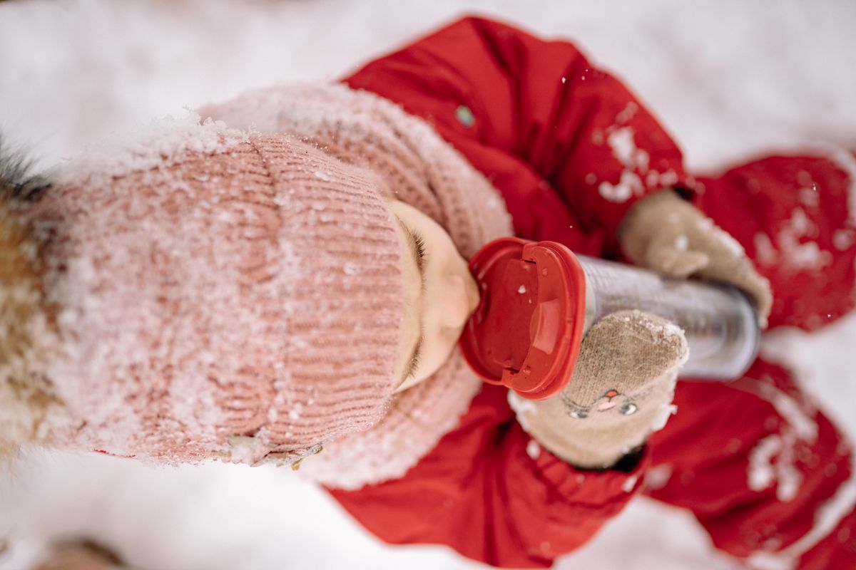 A child in snow-proof clothes enjoys a drink from a tumbler in a snowy outdoor setting.