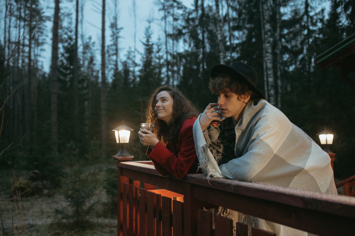 Two young adults enjoy a cozy evening on a forest deck, wrapped in blankets, sipping drinks.