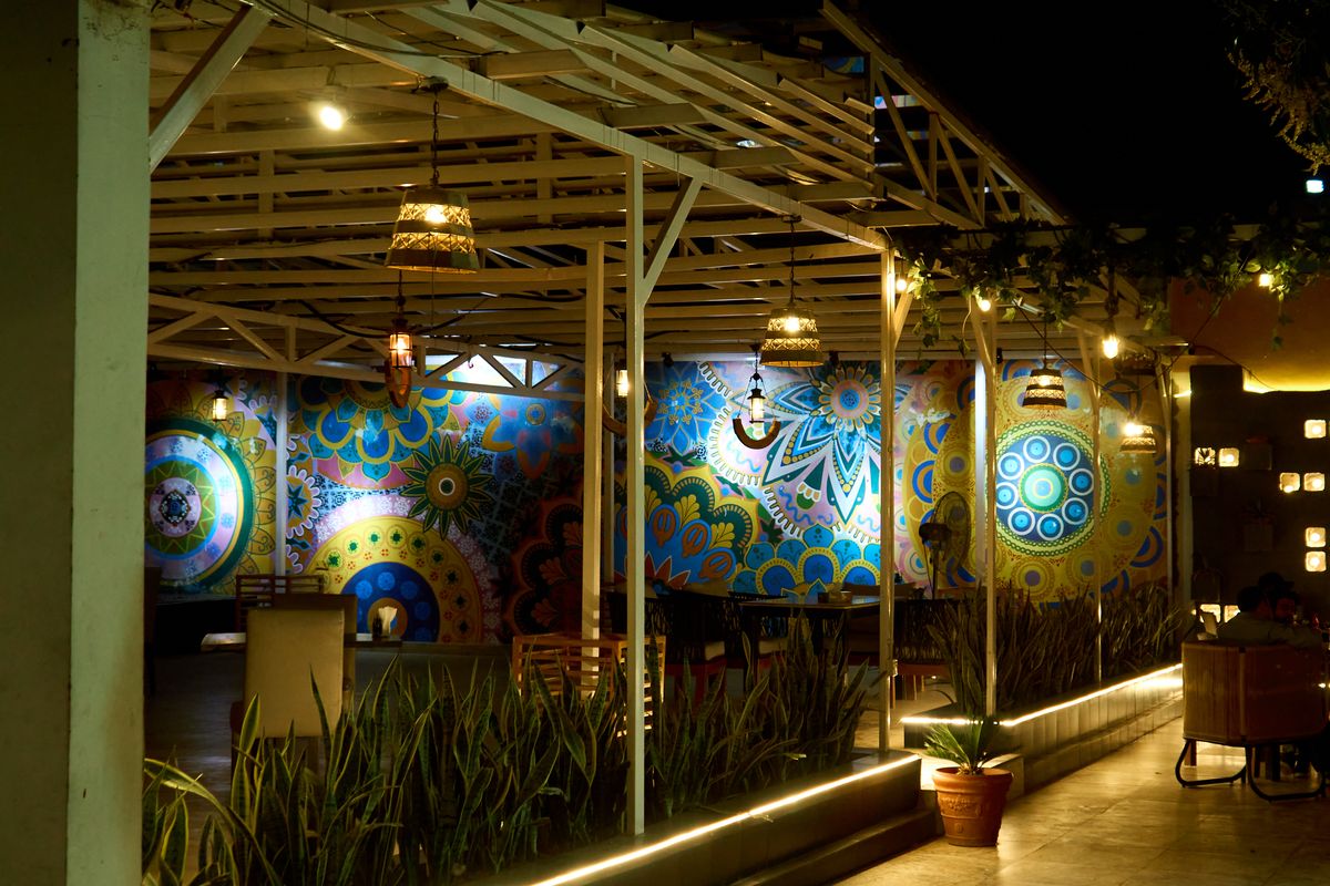 A vibrant outdoor patio with colorful wall murals illuminated at night, featuring intricate patterns and warm lighting.