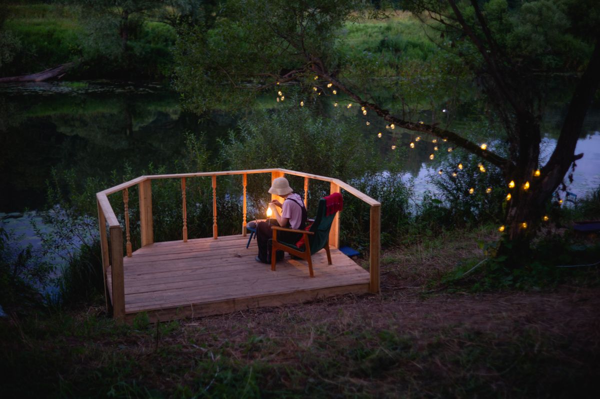 A serene evening scene with someone reading on a wooden deck by a river, lit by string lights.