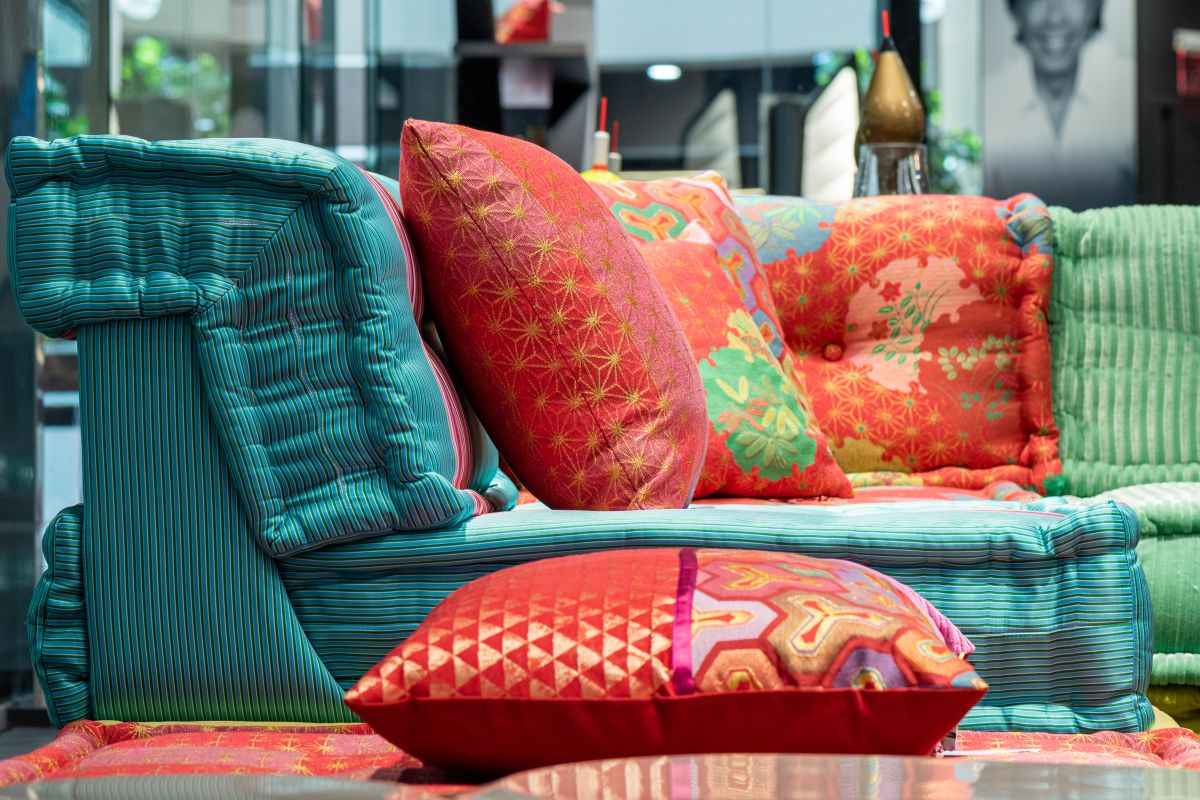 Comfortable bright sofa with abundance of soft colorful cushions placed near table with pillow in modern furniture store with glass wall