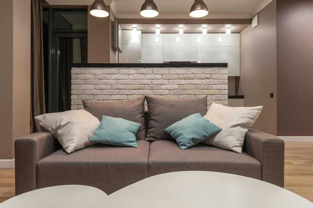 Comfortable gray sofa with colorful pillows placed near white table in stylish living room with glowing lights in modern apartment
