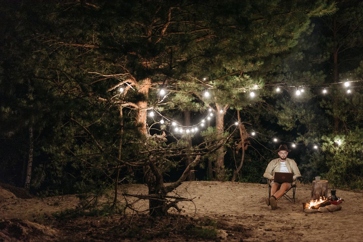 A man enjoys remote work on his laptop under string lights in a tranquil forest campsite at night.