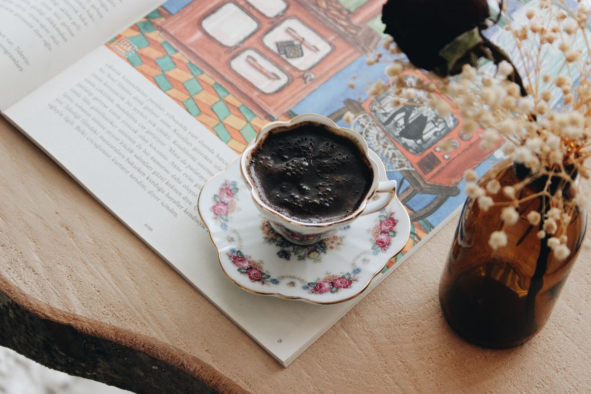A beautifully crafted cup of Turkish coffee on a floral porcelain saucer by a book and dried flowers.