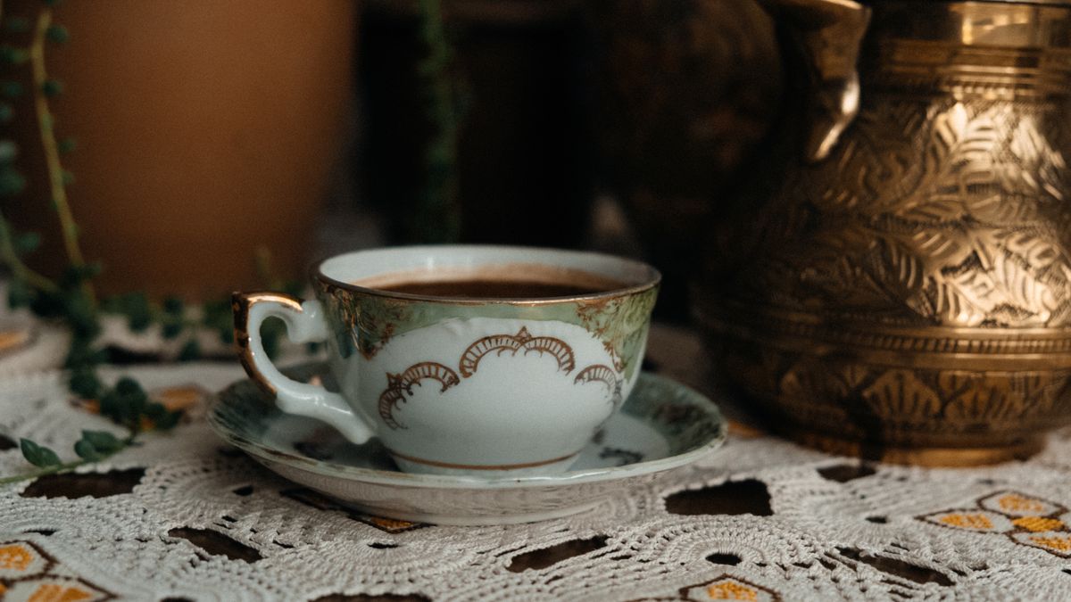 Elegant vintage porcelain cup filled with coffee on a lace tablecloth with brass pot in background.
