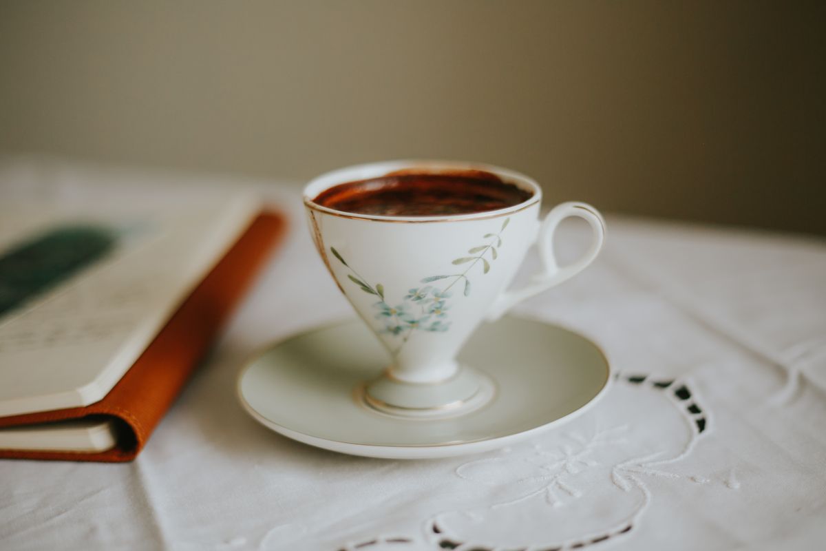 A floral porcelain teacup filled with coffee sits on a table beside a notebook, creating a calm, vintage vibe.