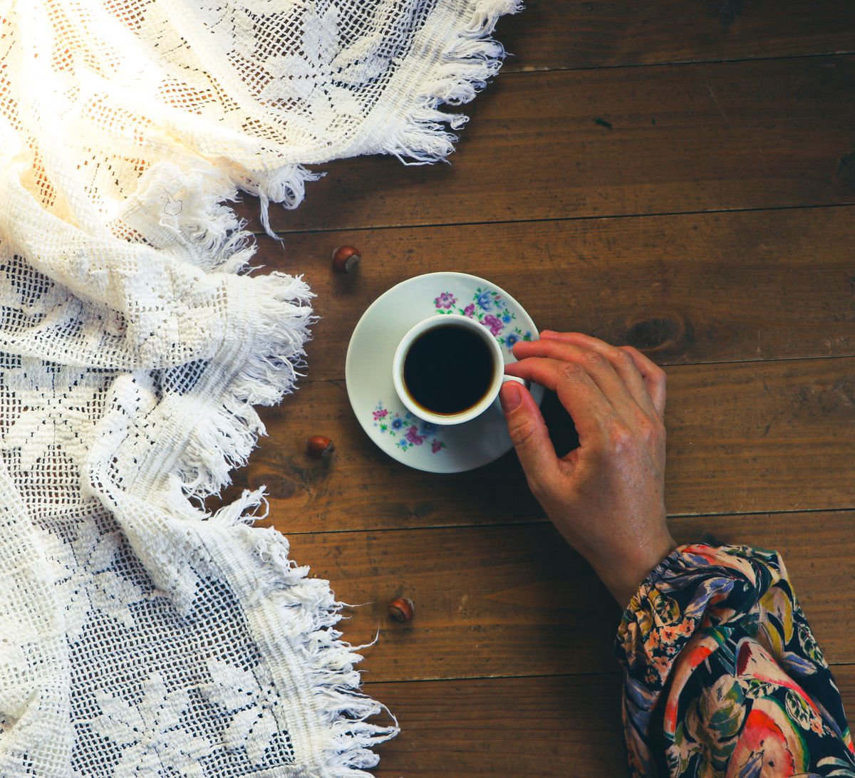 A single hand reaches for a coffee cup atop a lace-covered wooden table.