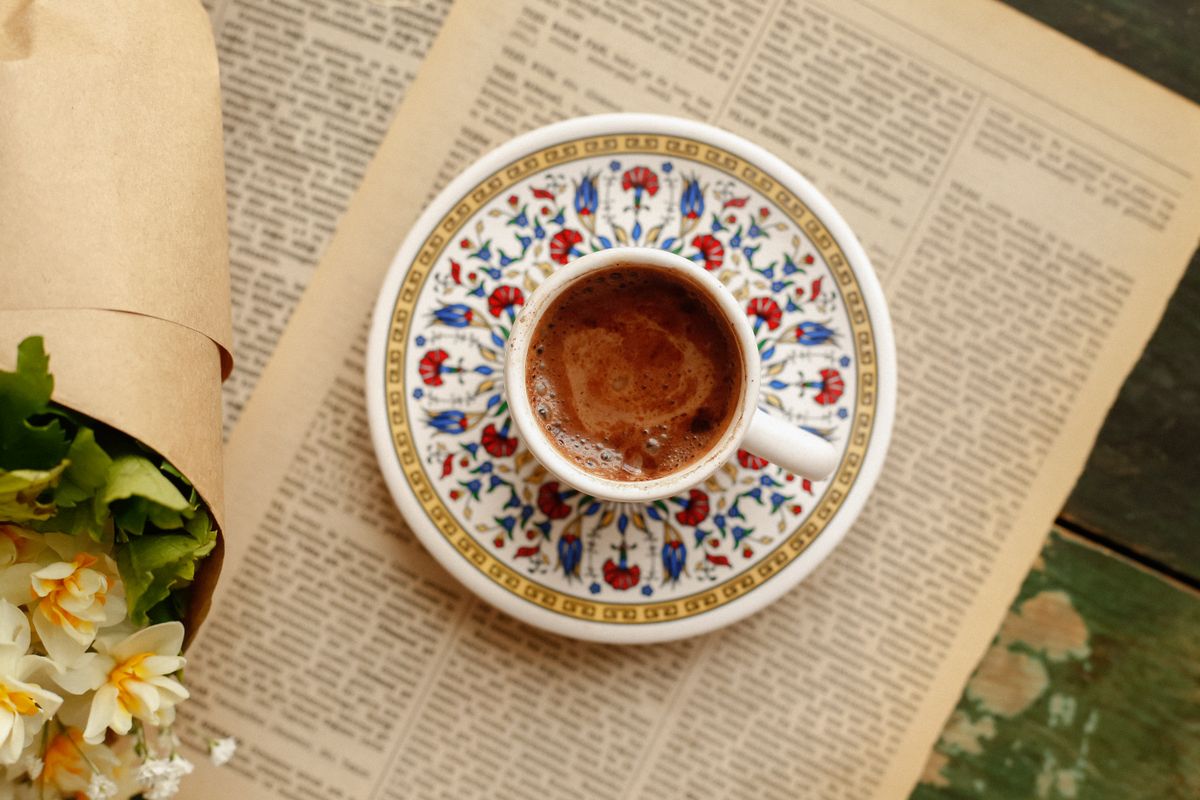 Elegant coffee cup with floral saucer atop a vintage newspaper beside a bouquet of flowers.