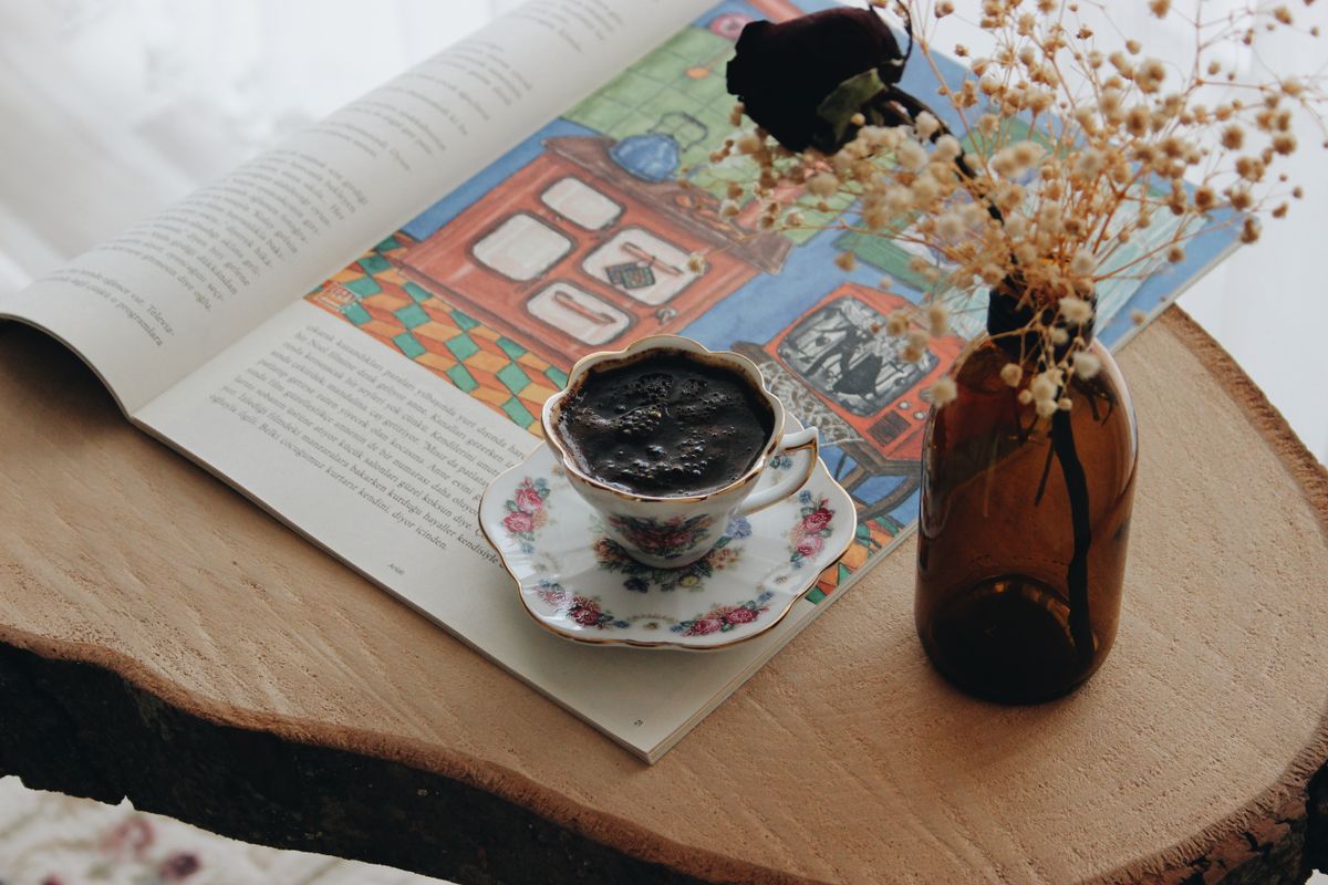 A vintage floral teacup filled with coffee sits on a magazine beside a vase of flowers on a wooden table.