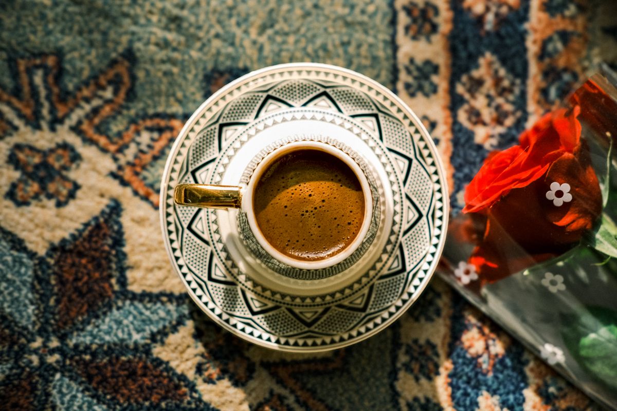 A top-down view of a Turkish coffee cup on a patterned rug with a red rose nearby.