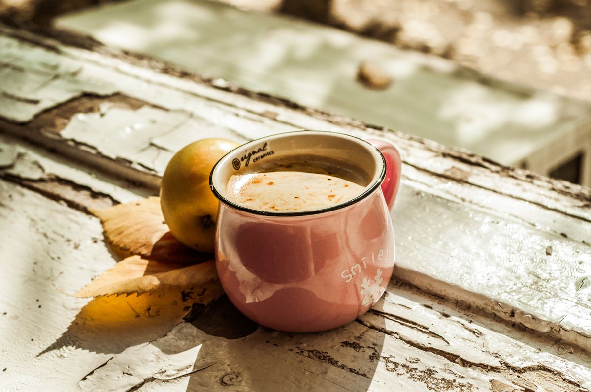 A pink ceramic mug of coffee on a rustic table with autumn leaves and an apple.