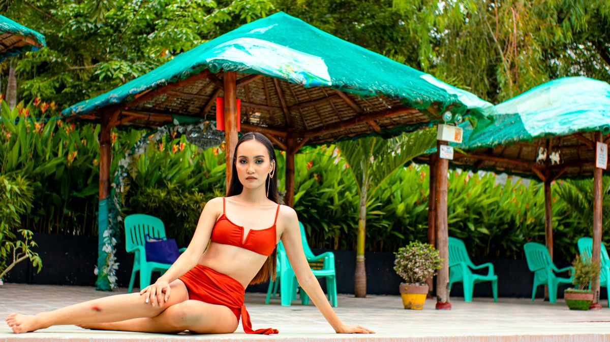 Asian woman in a red bikini reclining at a tropical outdoor resort under green-roofed huts.