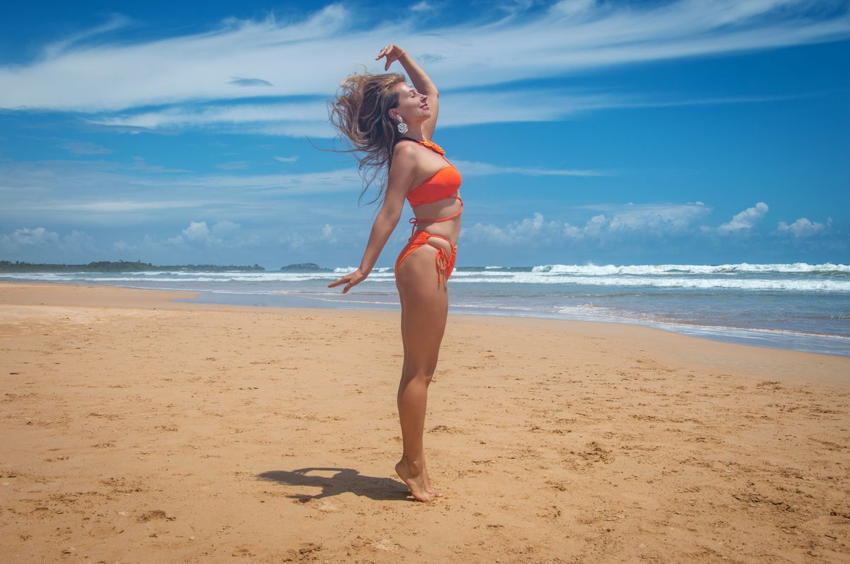 A woman in an orange bikini enjoying a sunny day on a sandy beach with blue sky.
