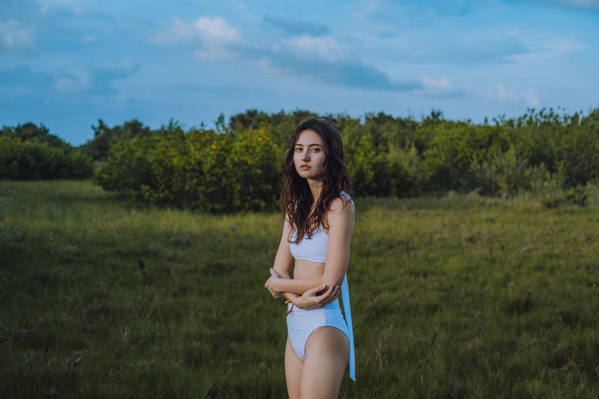Brunette woman in a white swimsuit standing in a grassy outdoor setting under a blue sky.