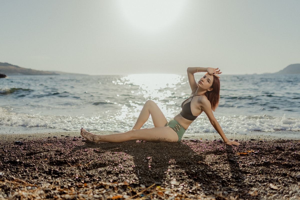 A woman in swimwear poses on a sandy beach with a serene ocean backdrop.