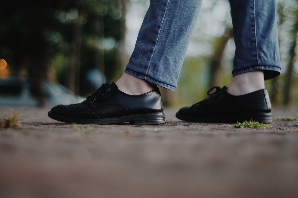 Side view of a person walking in black leather shoes and denim pants on a sidewalk.