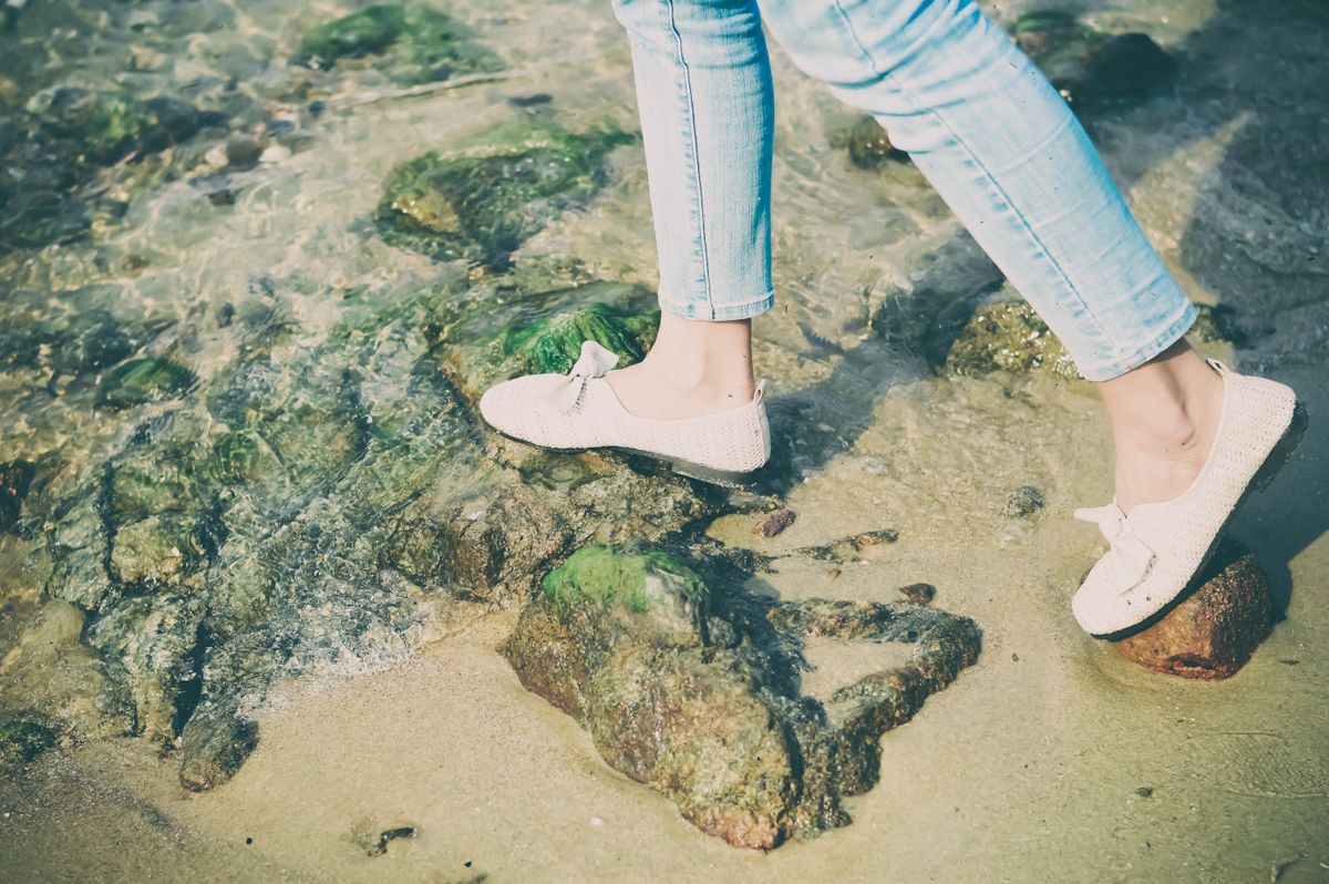 Person in casual shoes walking on rocky beach shore with clear water.