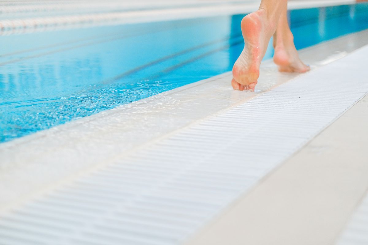 Close-up of bare feet walking on the wet edge of a swimming pool with clear blue water.