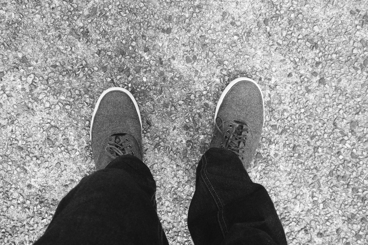 Monochrome image of feet wearing casual sneakers standing on gravel surface.