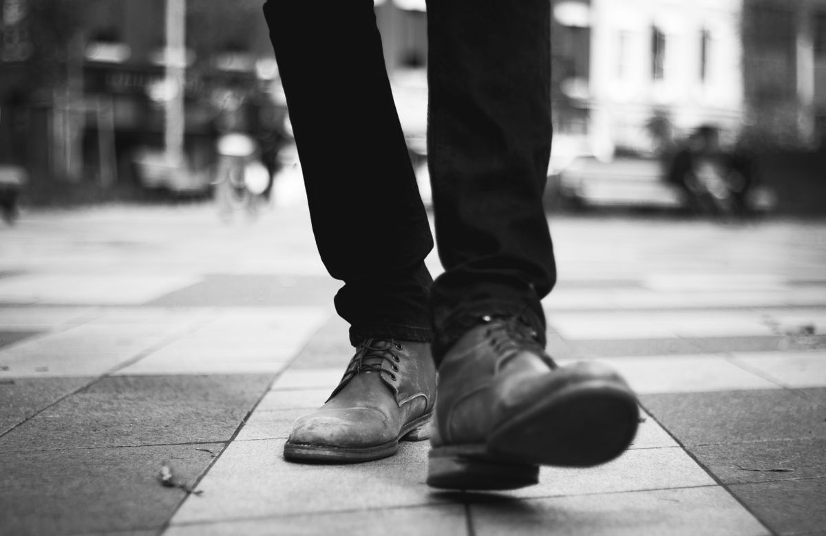Grayscale image of a man in boots walking on a tiled urban pavement.