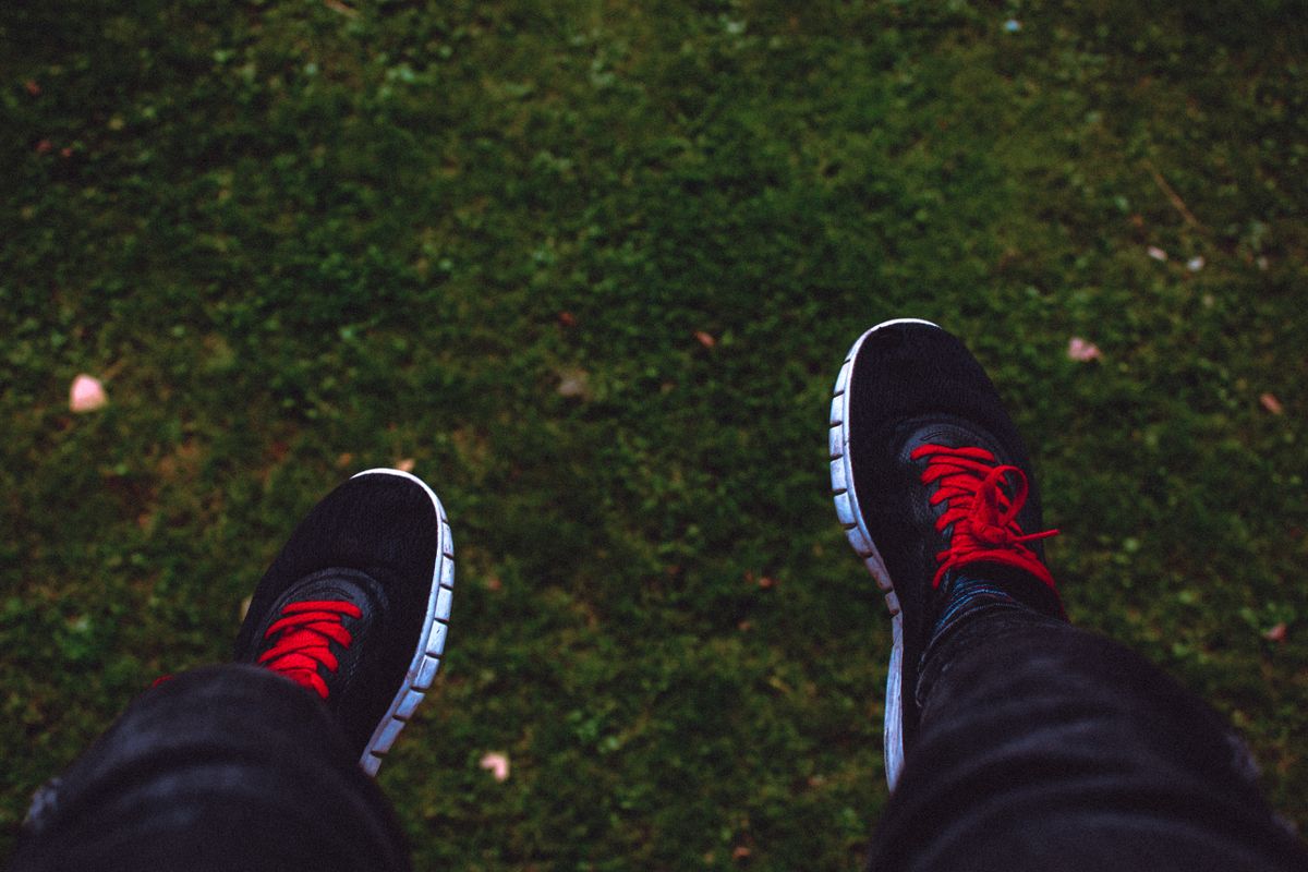 View of feet in black sneakers with red laces hovering above green grass.