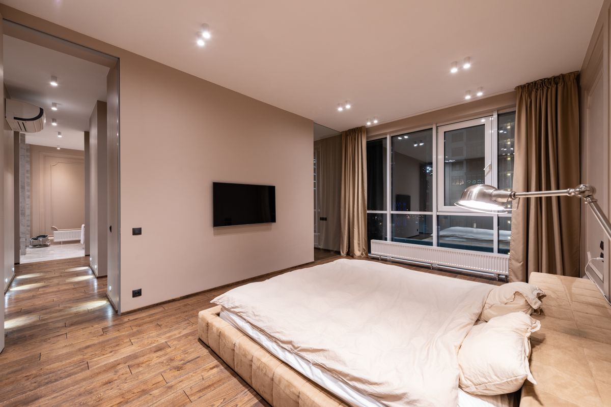 Creative design of bedroom with bed against TV and corridor with air conditioning above parquet in light house