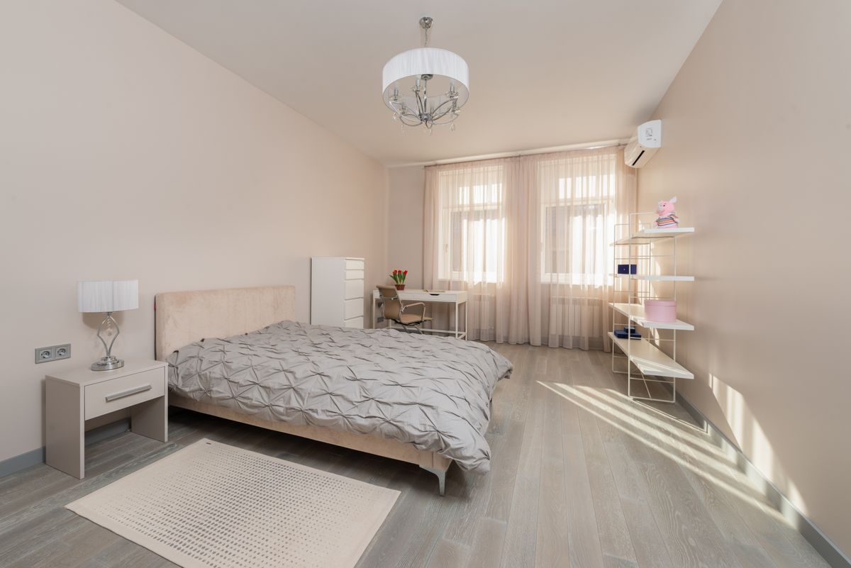 Comfortable bed with blanket placed near bedside table with lamp in spacious bedroom with table and shelves near window in apartment