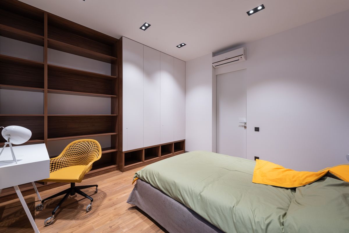 Interior of light room with bed and table with lamp and chair near white wall and cabinet with shelves on wooden floor