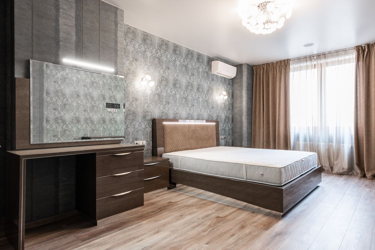 Comfortable bed with white mattress placed at wall near cupboard with mirror in modern spacious bedroom with window and curtains