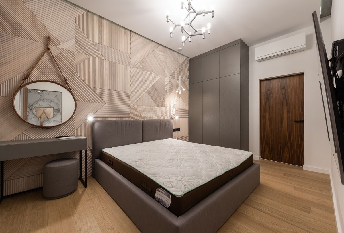Interior of modern light hotel room with bed near round mirror on wall near table and pouf near cupboard and door